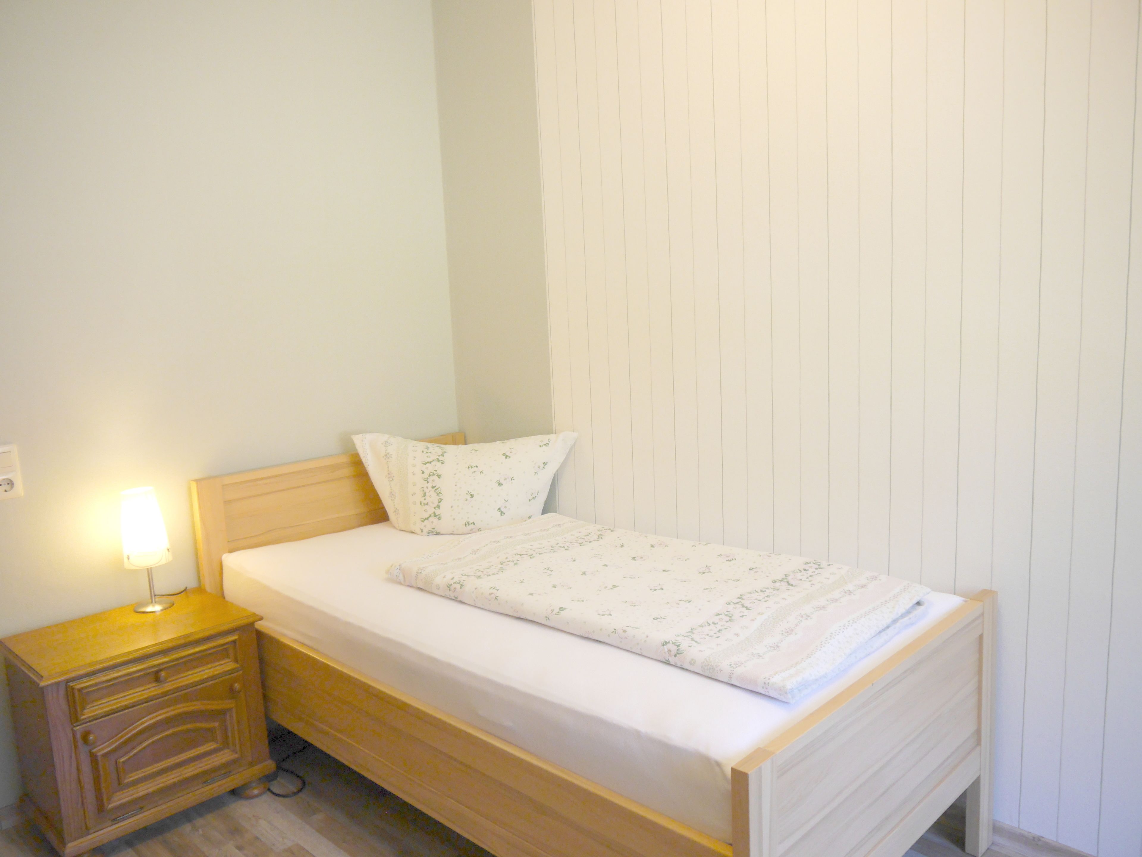 Una habitación pequeña con una cama individual, cabecera de madera, mesita de noche con lámpara y pared blanca lisa.