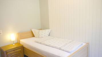 Una habitación pequeña con una cama individual, cabecera de madera, mesita de noche con lámpara y pared blanca lisa.