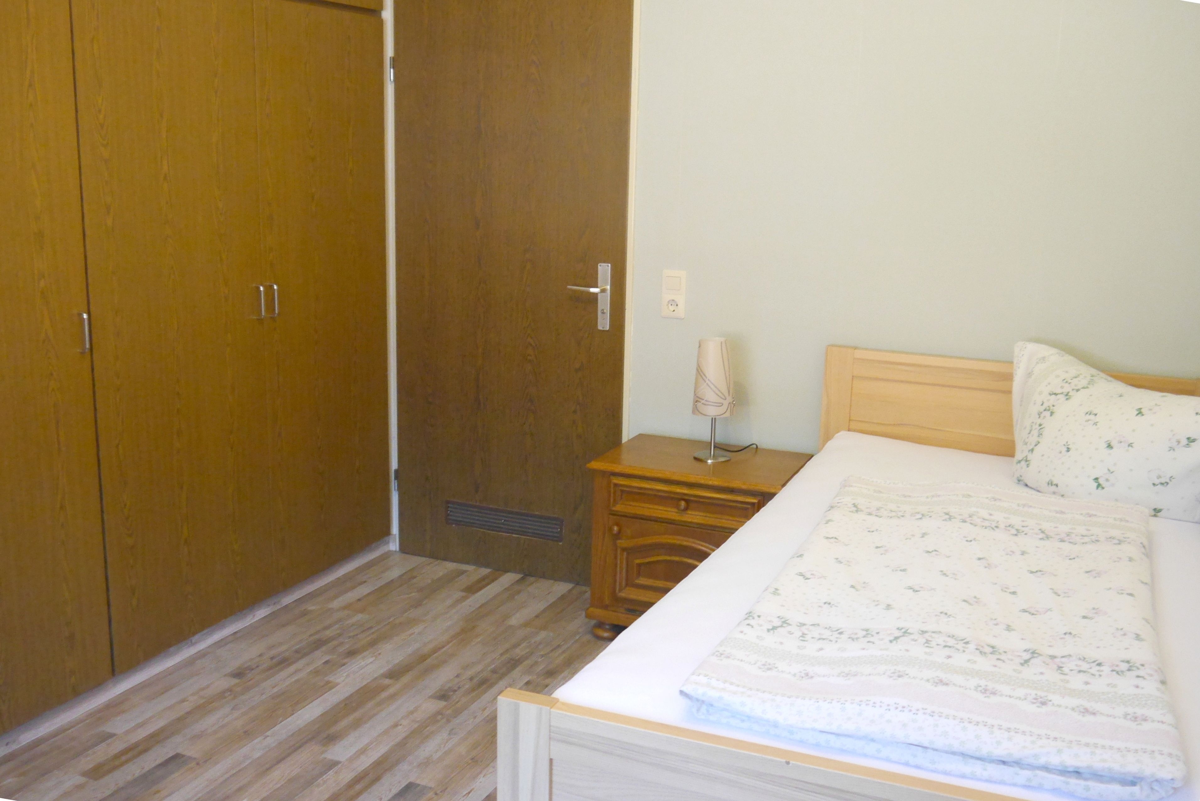 A bedroom with a wooden wardrobe, a bedside table with a lamp, and a bed with a floral-patterned blanket.