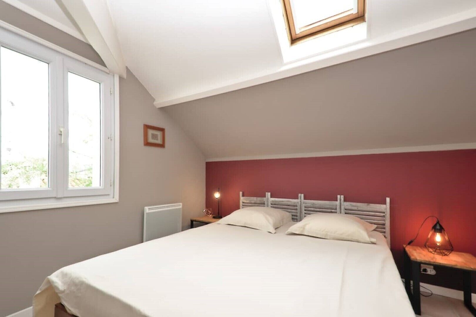 A bedroom with a bed, two bedside tables, a radiator, a window, and a skylight.