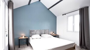 Une chambre avec un plafond en pente, un lit avec une literie blanche, deux tables de chevet avec des lampes et une fenêtre avec des rideaux.