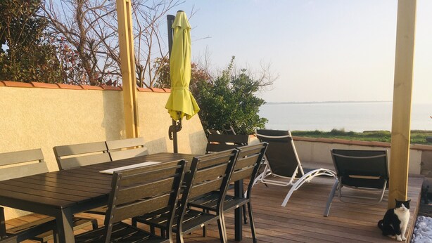 Une terrasse extérieure avec une plateforme en bois, une table et des chaises, un parasol jaune et une vue sur l’eau.