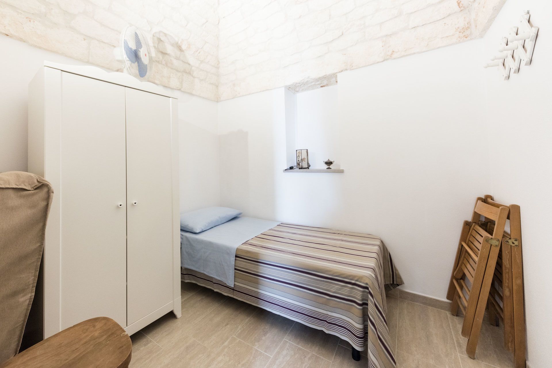 A small, well-lit bedroom with a single bed, a wardrobe, a small shelf with decorative items, and a wooden chair.
