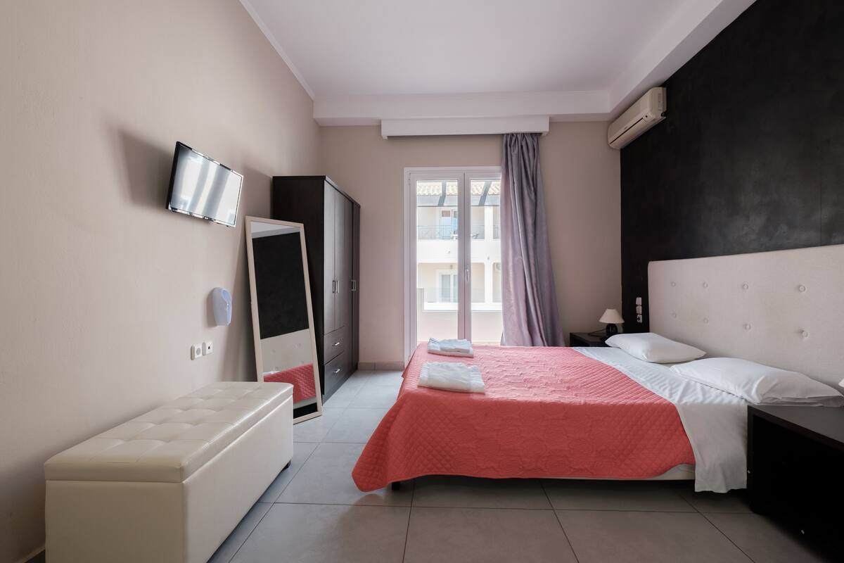 Una habitación de hotel con cama, un banco, televisor, armario y ventana con cortinas.