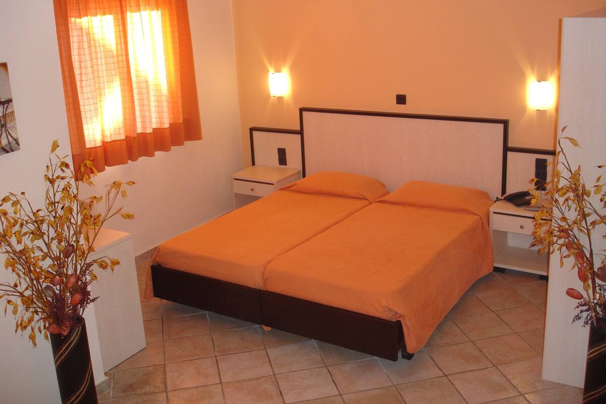 Una cama doble con estructura de madera, dos mesitas de noche, una lámpara de noche, una ventana con cortinas anaranjadas y dos luces fijadas a la pared.