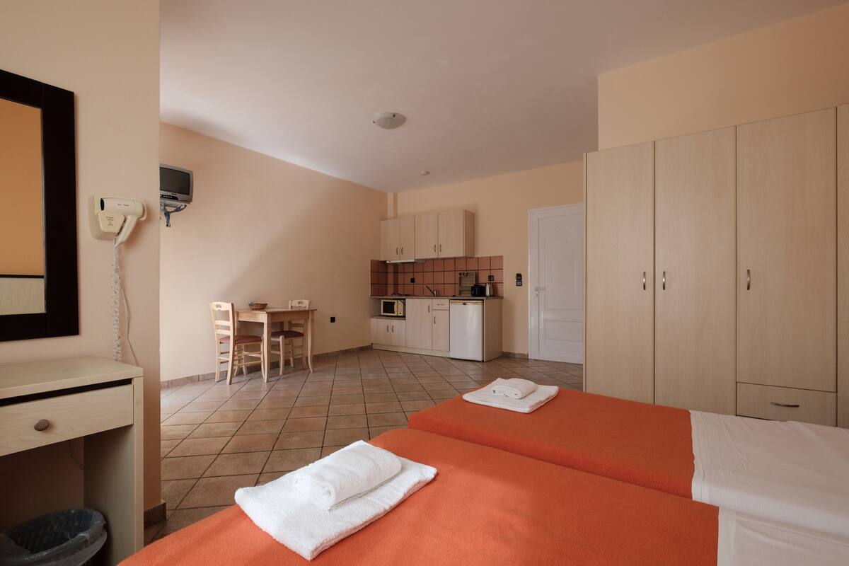 Habitación de hotel con dos camas, un escritorio, una silla, una kitchenette pequeña y un armario.