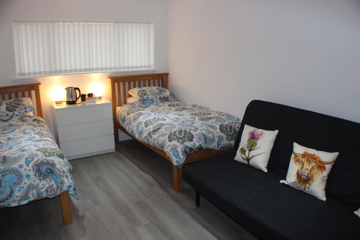 A bedroom with two single beds, a sofa, a bedside table with a lamp, and a window with blinds.
