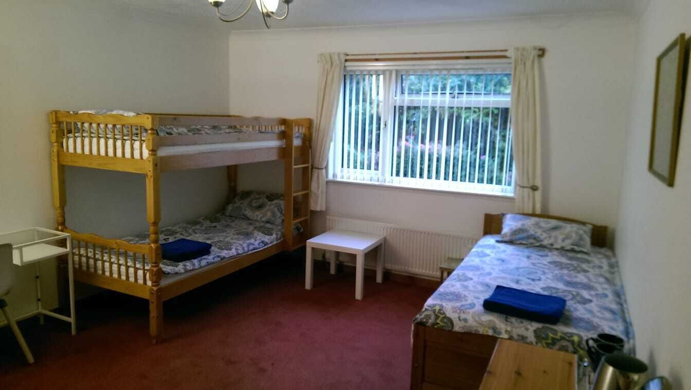 A dormitory room with two bunk beds, a small desk, a radiator, and a window with curtains.