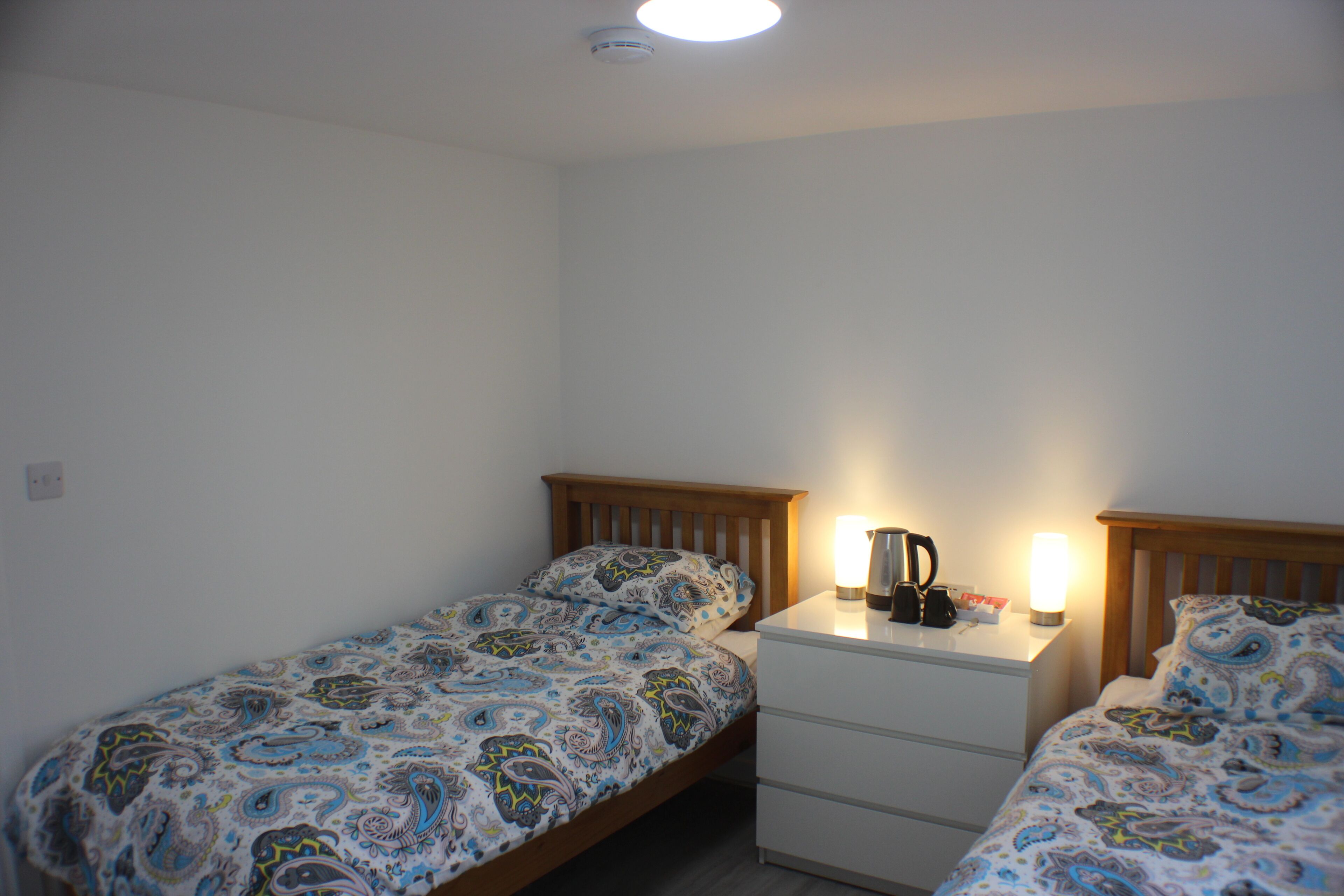 Quarto duplo com duas camas de solteiro, uma mesinha de cabeceira com abajur e um criado-mudo com cafeteira.