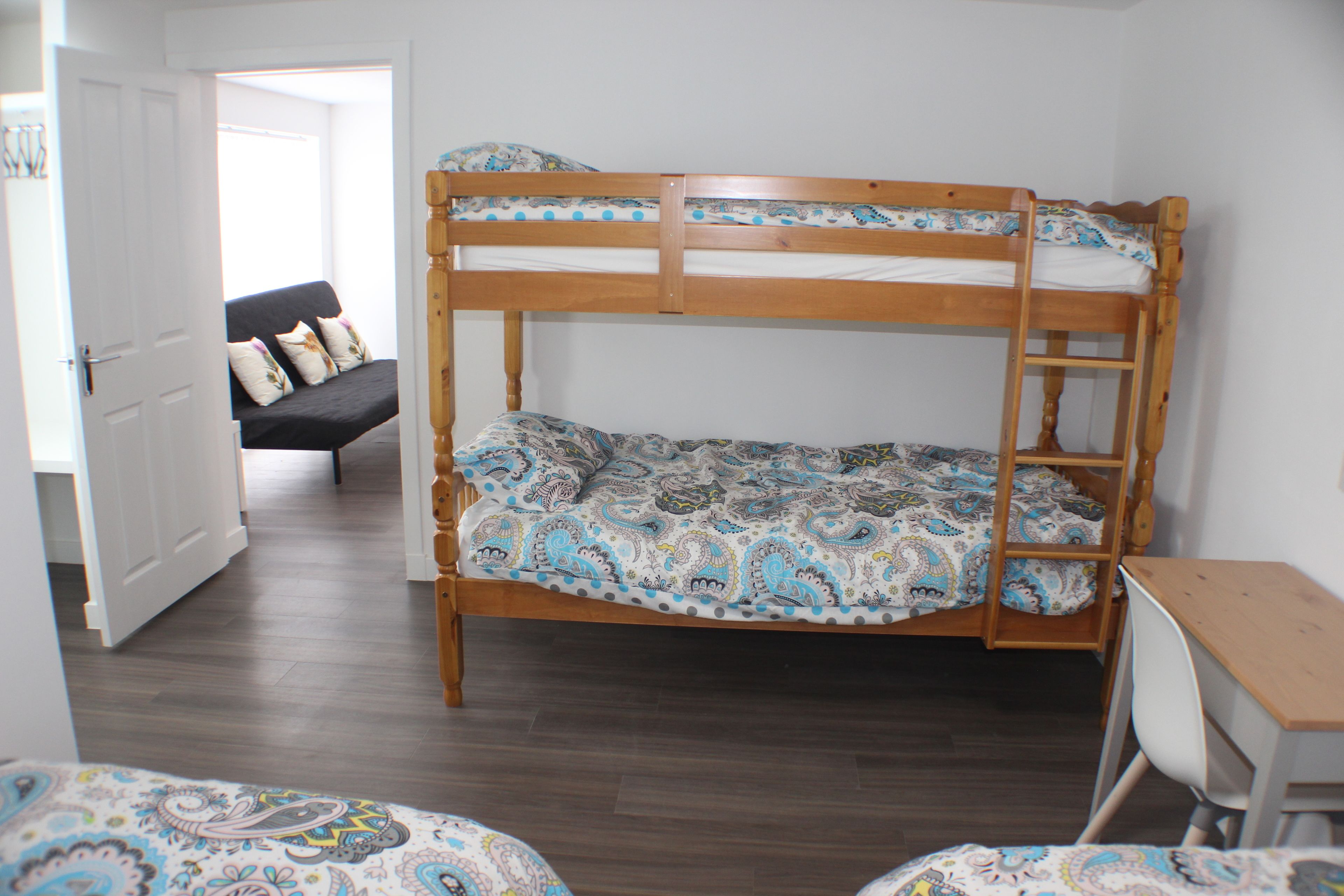 Una cama individual superpuesta con colcha estampada, un escritorio con silla y un sofá en una habitación con suelo de madera y paredes blancas.