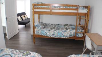 Una cama individual superpuesta con colcha estampada, un escritorio con silla y un sofá en una habitación con suelo de madera y paredes blancas.