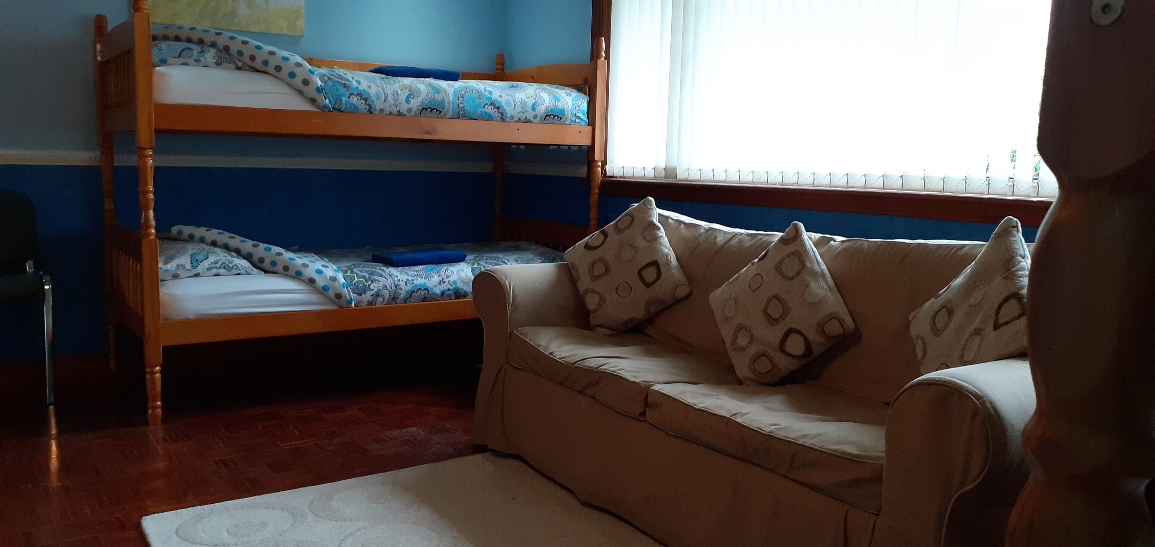 Una cama individual con ropa de cama azul y blanca, un sofá color beige con cojines estampados y una ventana con persianas.