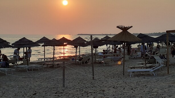 A beach with sun loungers and umbrellas, a calm sea, and a sunset in the background.