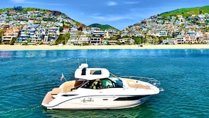 Un yacht baptisé « Aperitif » navigue sur une eau d’un bleu limpide, avec en arrière-plan une zone résidentielle densément peuplée, s’étendant sur une colline.