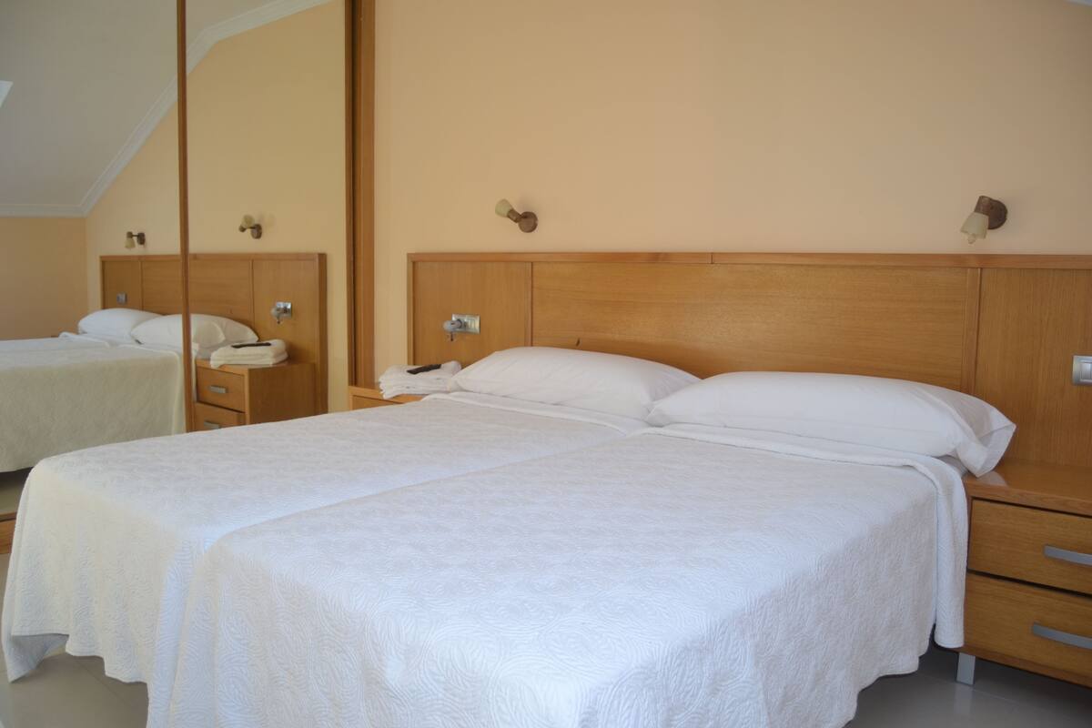 A hotel room with two beds, wooden headboards, bedside tables, and wall-mounted lamps.
