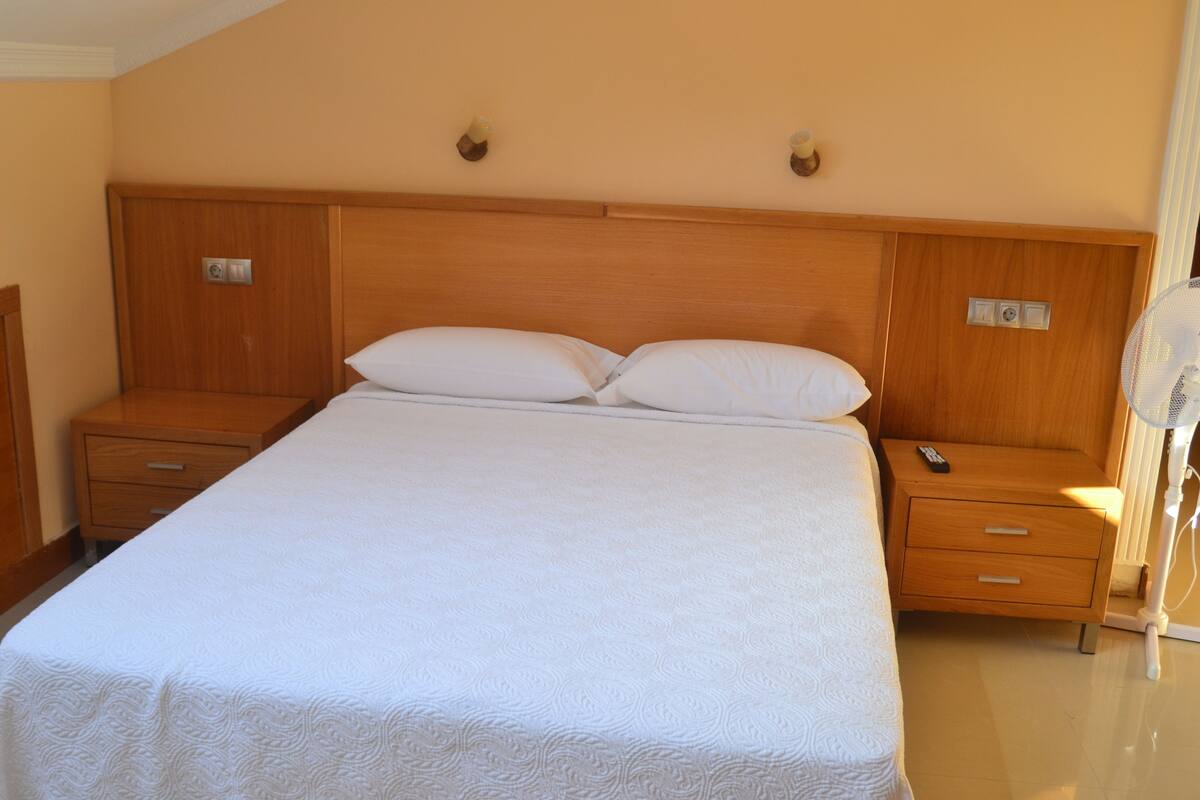 A large bed with a wooden headboard, two bedside tables with drawers, and a remote control on one of the tables.