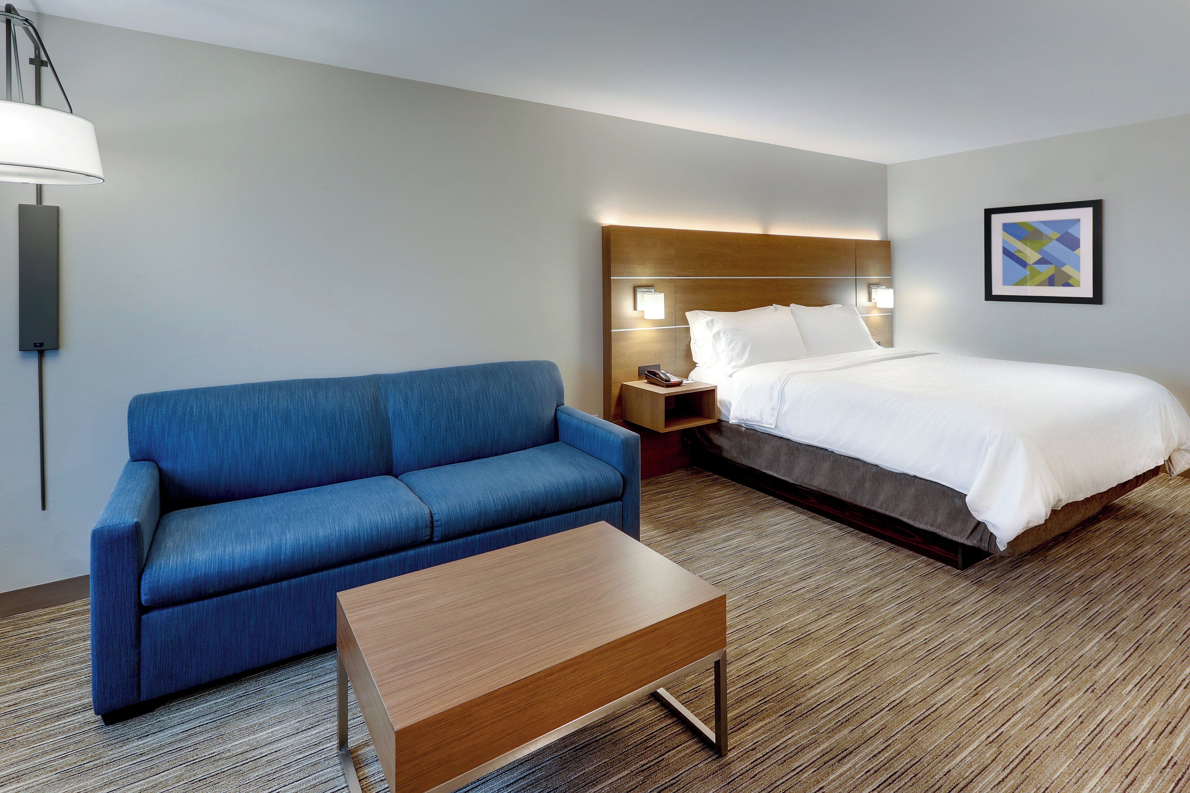Een hotelkamer met een blauwe bank, een houten salontafel, een bed met wit beddengoed, een nachtkastje met een lamp en een lijst met een afbeelding aan de muur.