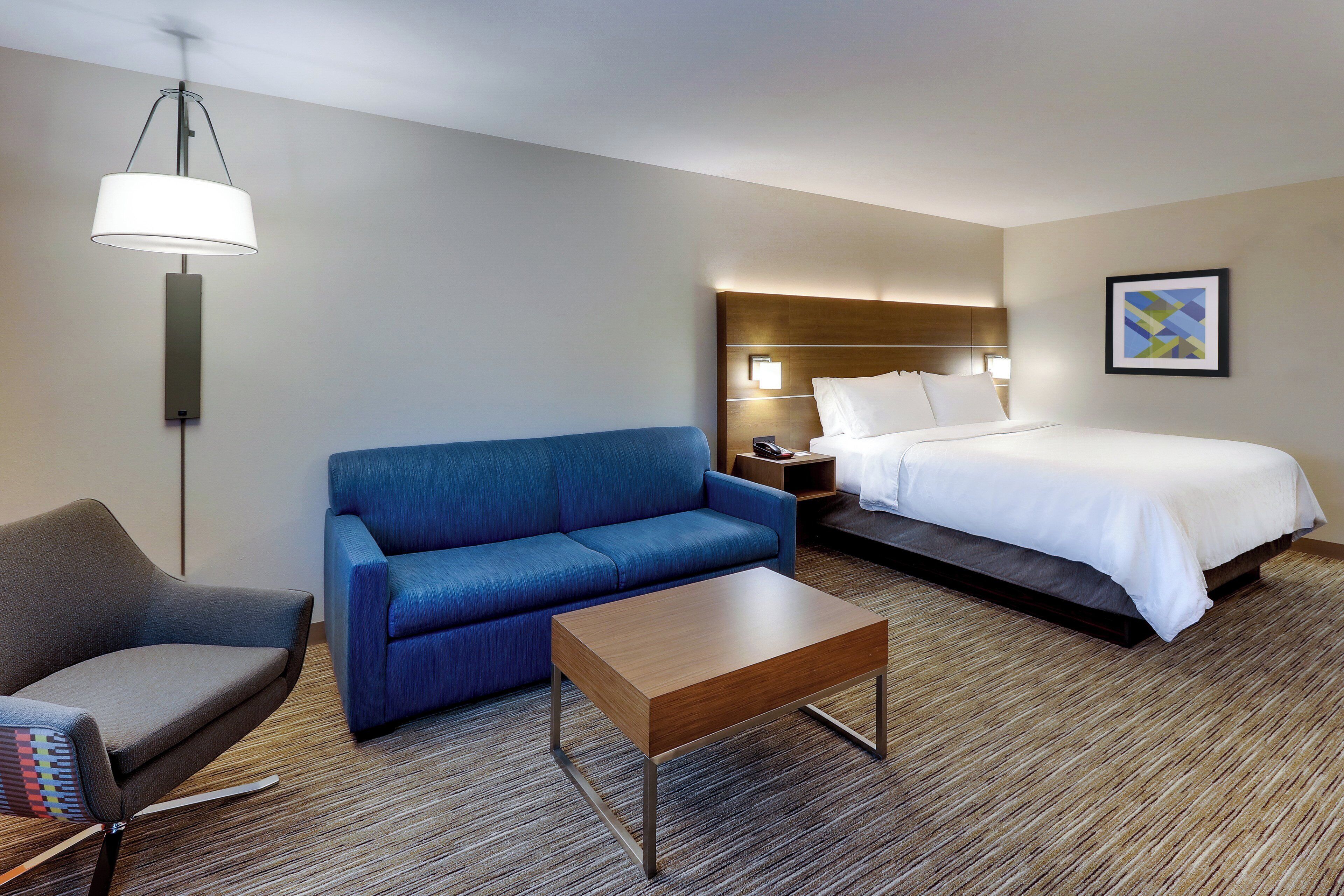 Una habitación de hotel con una cama, un sofá azul, una mesa de centro de madera, un sillón gris y una lámpara de pie.