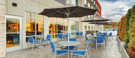 An outdoor patio with tables and chairs, a building with large windows, and a car parked nearby.