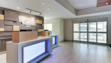 A hotel reception area with a modern design, featuring a reception desk, a digital display, and a view through the glass doors.