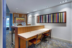 A modern reception area with a wooden desk, two chairs, a colorful striped artwork on the wall, and a patterned carpet. - Holiday Inn Express & Suites Middletown - Goshen by IHG (Middletown)