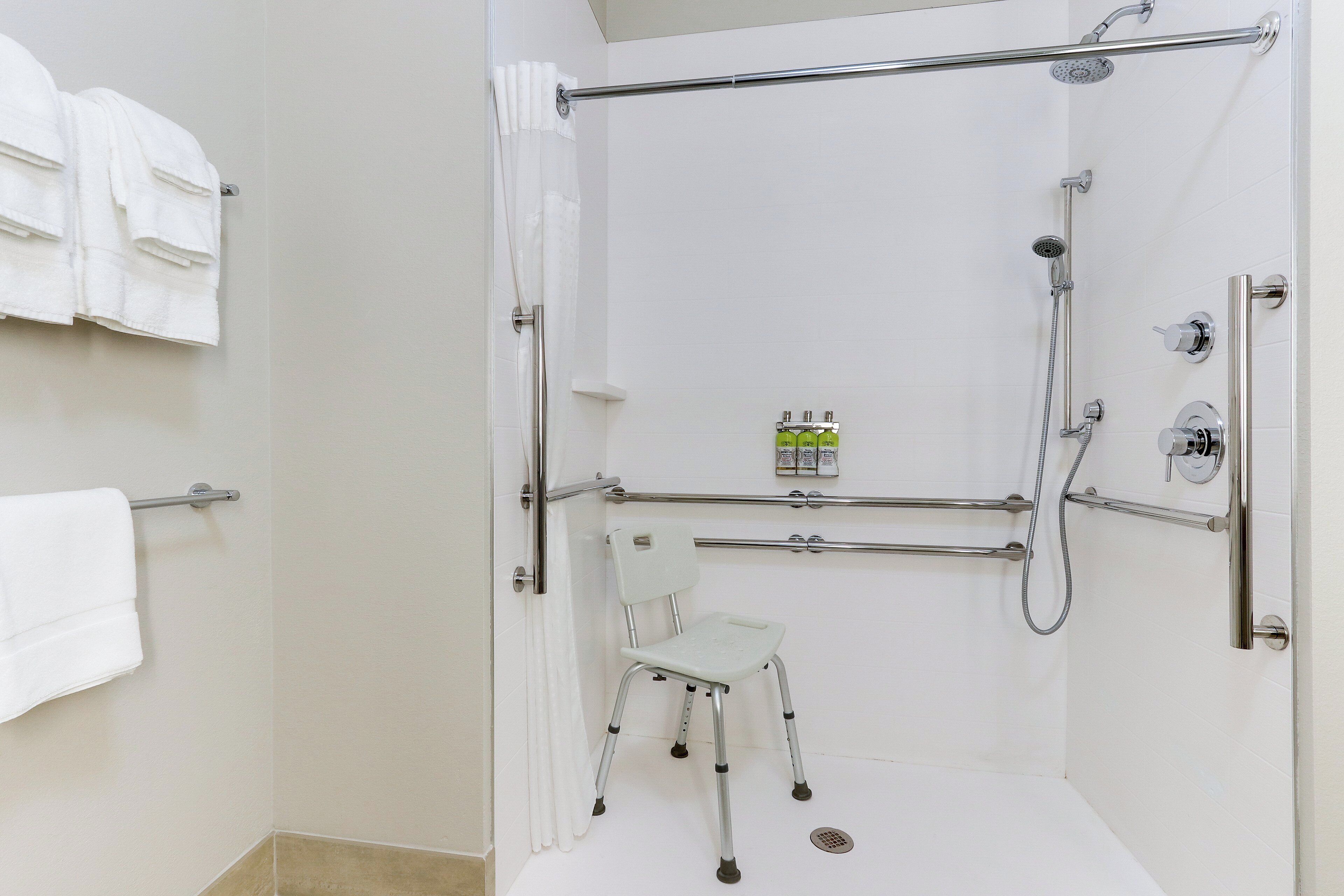 Une salle de bain moderne avec une douche à l’italienne, des barres d’appui, une chaise blanche et un porte-serviettes.