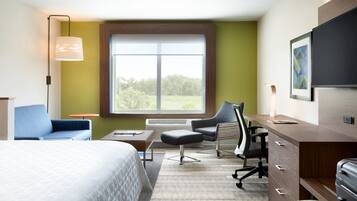 Habitación de hotel con cama, escritorio, silla, lámpara y ventana con vista a árboles.