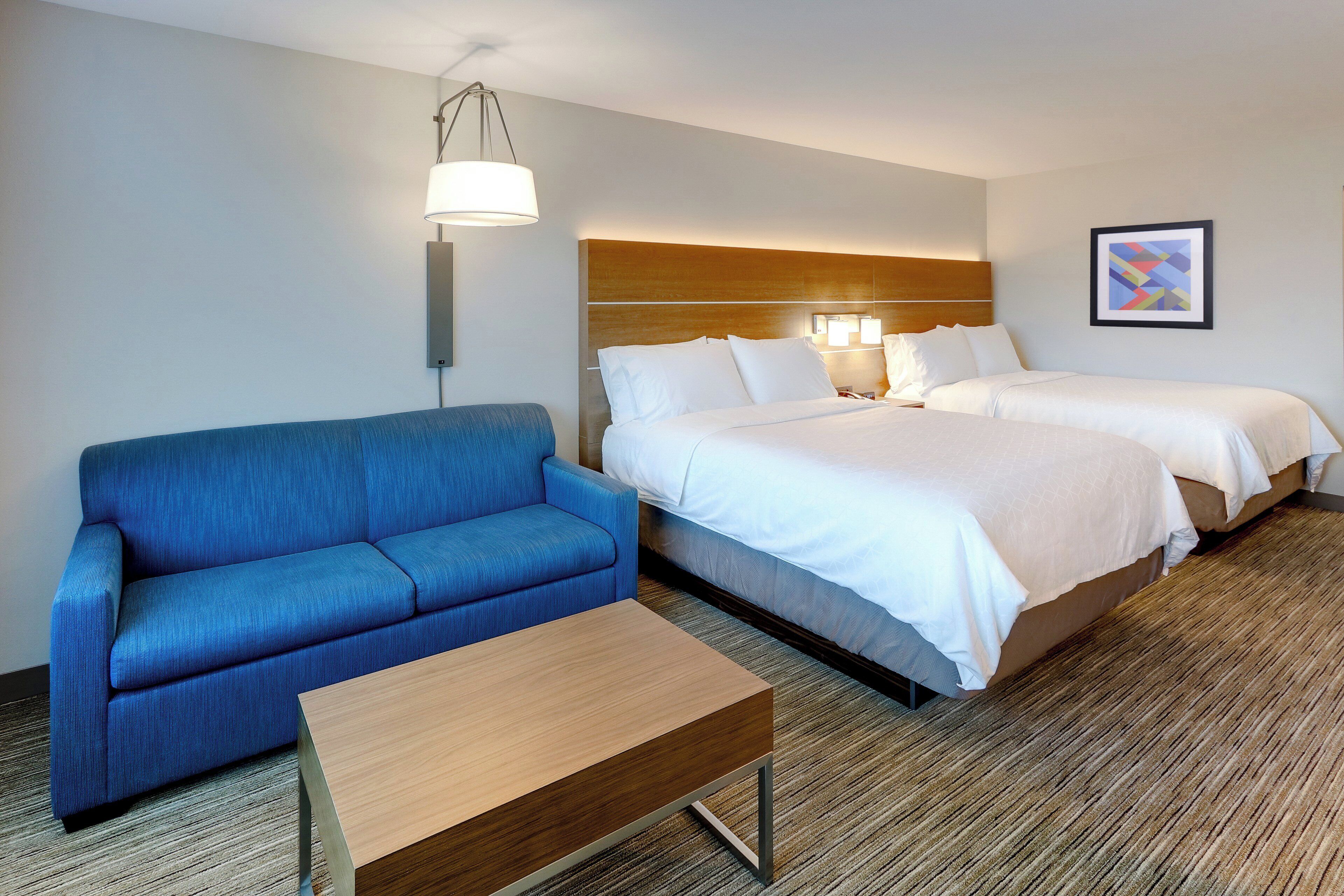 Een hotelkamer met een blauwe bank, een houten salontafel, twee bedden met witte lakens, een houten hoofdbord, een wandlamp en een ingelijst schilderij aan de muur.