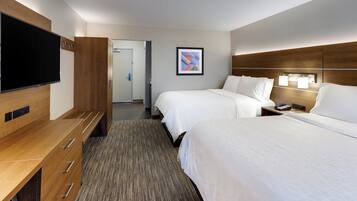 A hotel room with two beds, a TV, a desk with drawers, a bedside table with a lamp, and a door leading to a hallway.