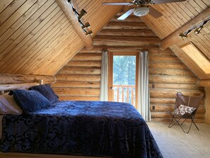 Cozy cabin bedroom with a sloped ceiling and wooden beam structure, featuring a bed with a patterned bedspread and a small chair with a checkered cushion.