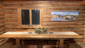 A log cabin interior with a wooden dining table, two cushioned benches, a window with a view of mountains, and a painting of a snowy landscape. - Taylor Canyon Cabin in Almont (Almont)