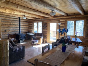 Cozy cabin interior with a central dining table, wood-burning stove, and large windows offering scenic forest views.