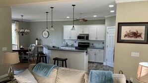Un soggiorno con un divano beige, un'area pranzo con un lampadario e una cucina con pensili bianchi e un forno a microonde.