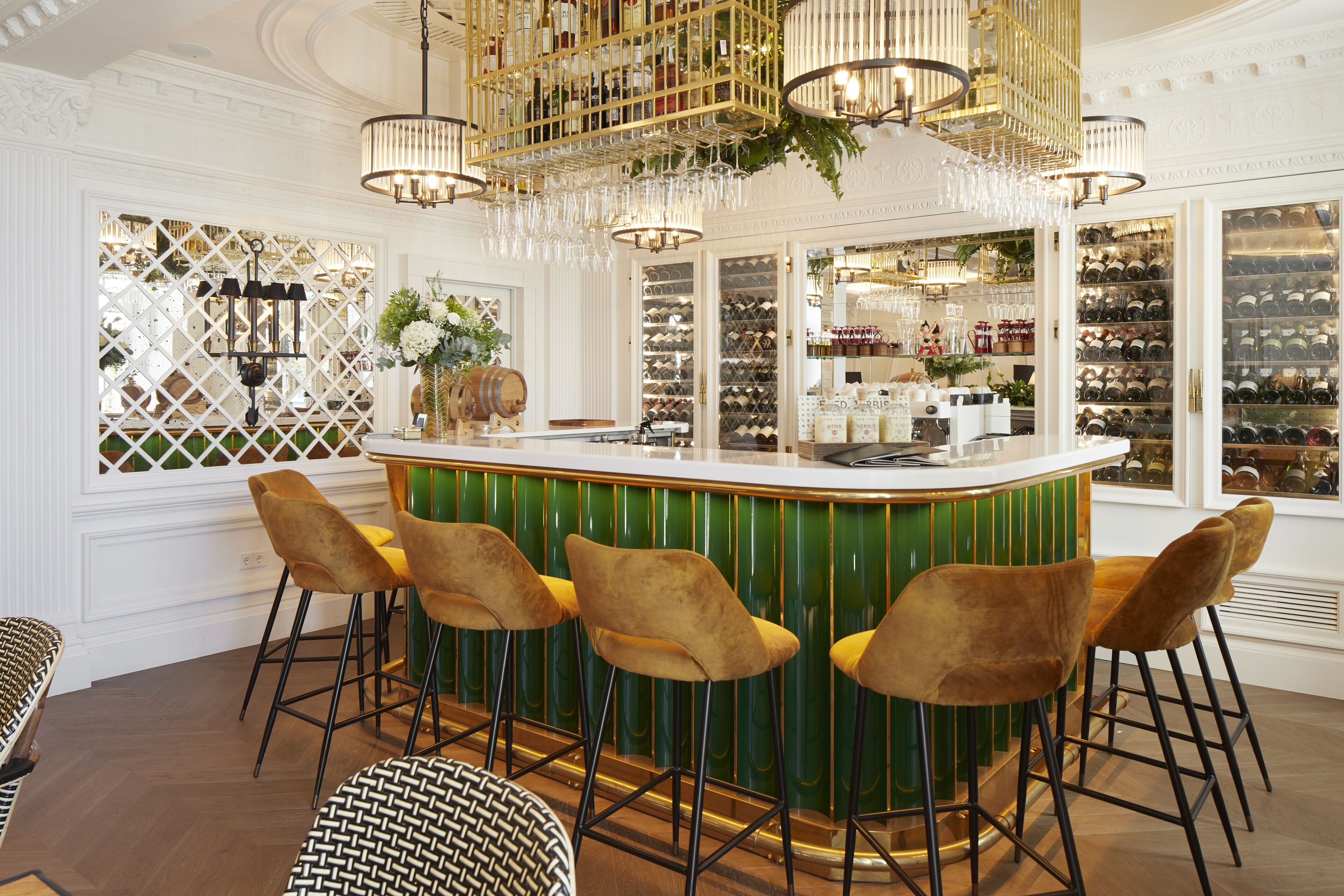 Uma área de bar com balcão verde, quatro banquetas douradas e uma adega ao fundo.