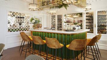 Uma área de bar com balcão verde, quatro banquetas douradas e uma adega ao fundo.