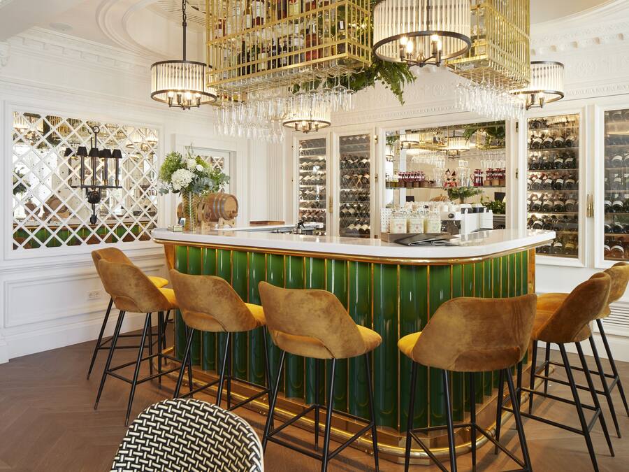 Uma área de bar com balcão verde, quatro banquetas douradas e uma adega ao fundo.