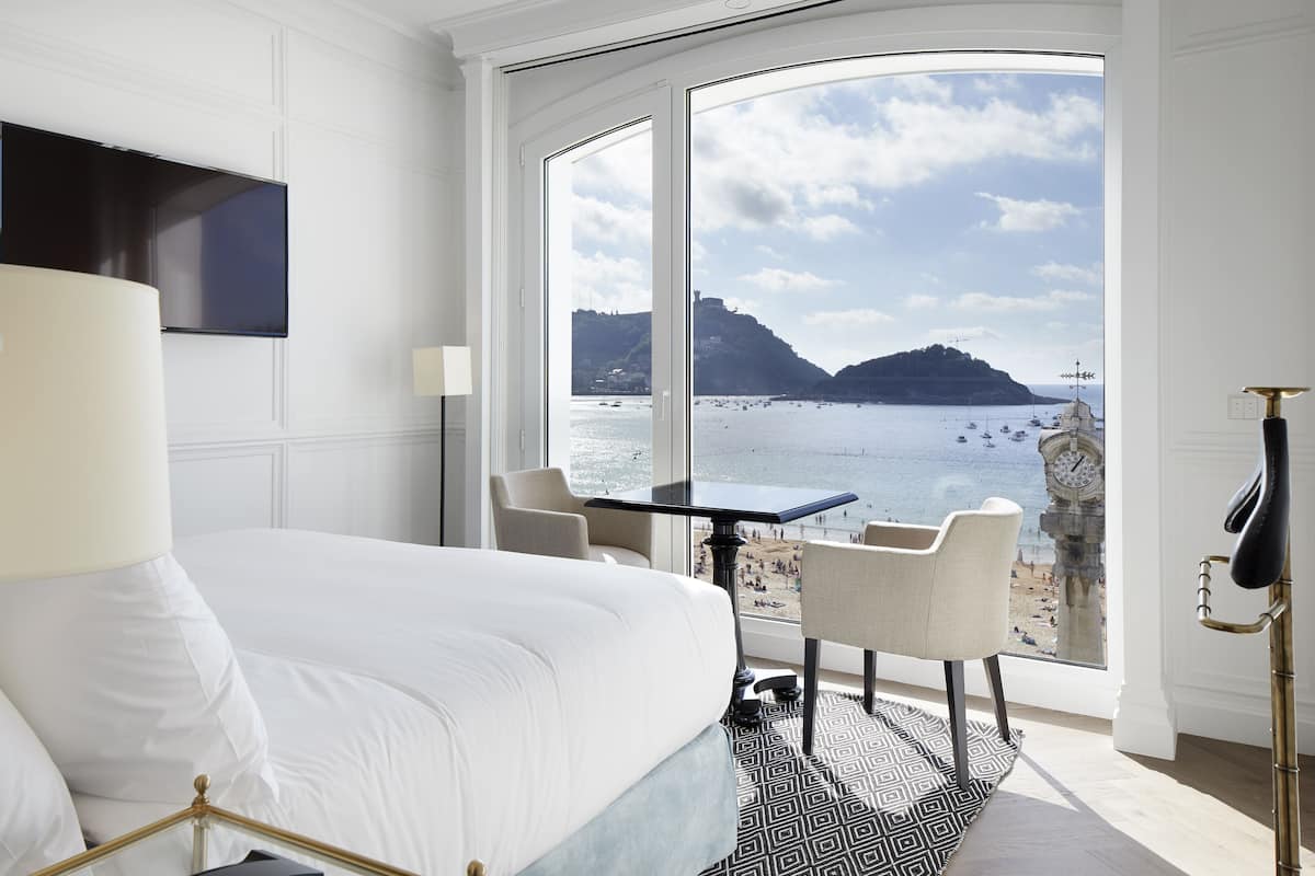 Una habitación de hotel con una cama, un televisor montado en la pared, una mesa con sillas y vistas al mar y un campanario.