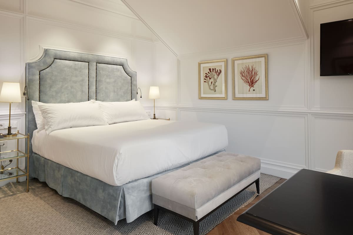 Habitación de hotel ordenada con una cama grande, lámparas de noche, una banca y un televisor de pantalla plana.