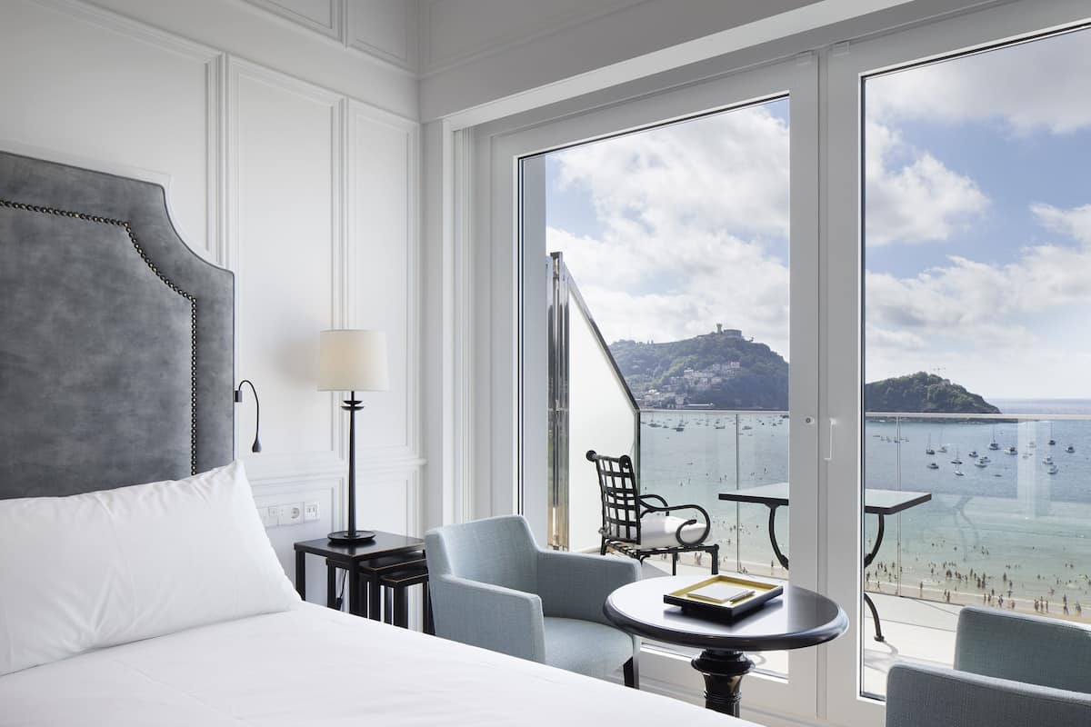 Una habitación de hotel con una cama grande, una zona de estar con una mesa, y un balcón con vista al mar y una isla distante.