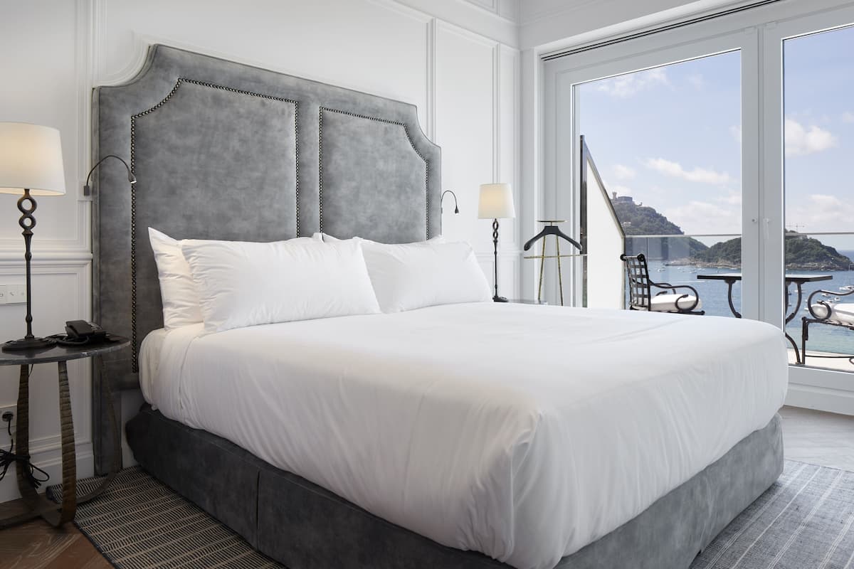 Una cama grande con cabecera tapizada en gris, ropa de cama blanca y vista al mar a través de amplios ventanales.