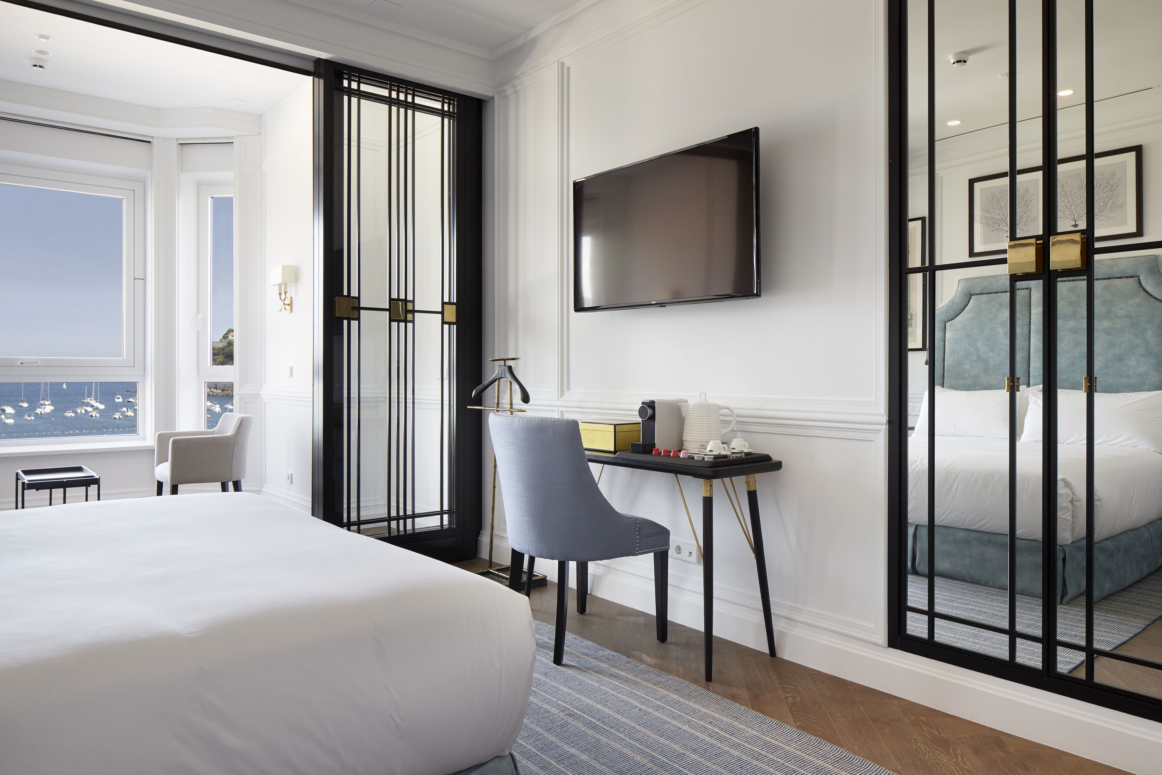 A hotel room with a large bed, a TV mounted on the wall, a desk with a chair, and a view of the sea through a window.