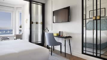A hotel room with a large bed, a TV mounted on the wall, a desk with a chair, and a view of the sea through a window.
