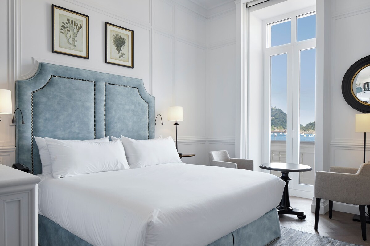 A large bed with a blue headboard, two bedside lamps, a round table, and a chair in a room with a large window and framed artwork on the wall.