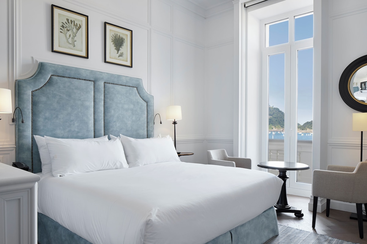 A large bed with a blue headboard, two bedside lamps, a round table, and a chair in a room with a large window and framed artwork on the wall.