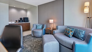 A modern hotel room with a grey sofa, two armchairs, a small table with a lamp, a kitchenette, and a floor lamp. - Courtyard by Marriott Bentonville Rogers Promenade (Rogers)