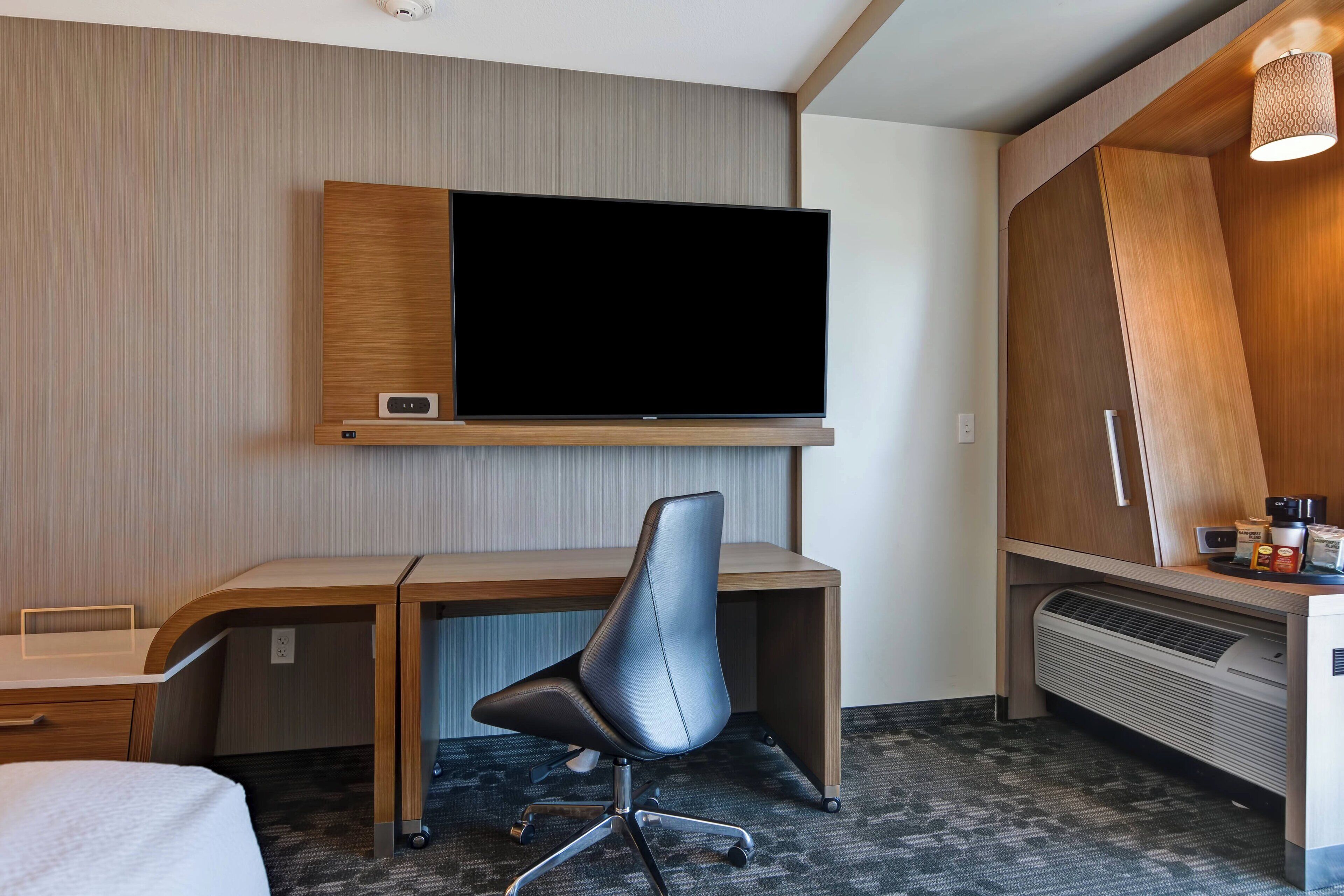 Une chambre d’hôtel avec un bureau en bois, une chaise moderne, un téléviseur à écran plat fixé au mur, un radiateur et une petite table sur laquelle se trouve une machine à café.