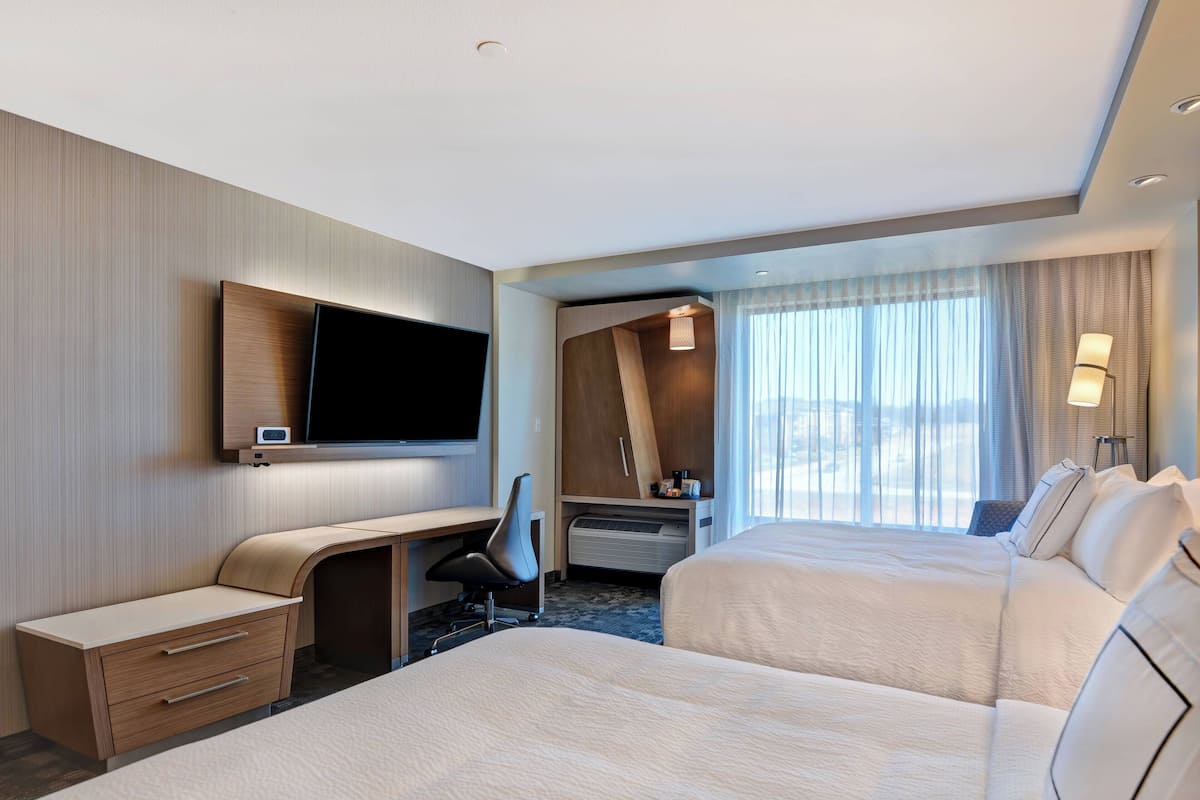 Une chambre d’hôtel avec deux lits, un bureau, une chaise, une télévision et une grande fenêtre avec des rideaux.