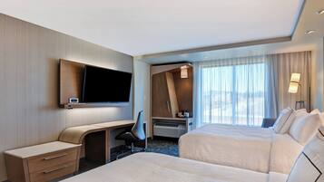 A hotel room with two beds, a desk, a chair, a TV, and a large window with curtains.