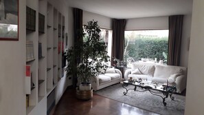 A living room with a white sofa, a coffee table, and a large potted plant. There is a bookshelf with books and decorative items on the left wall. - B&B Villa Victoria (Ragusa)