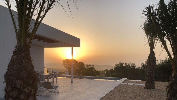 Un espace extérieur moderne, aménagé en terrasse, avec une piscine, des palmiers et une vue sur le coucher de soleil.